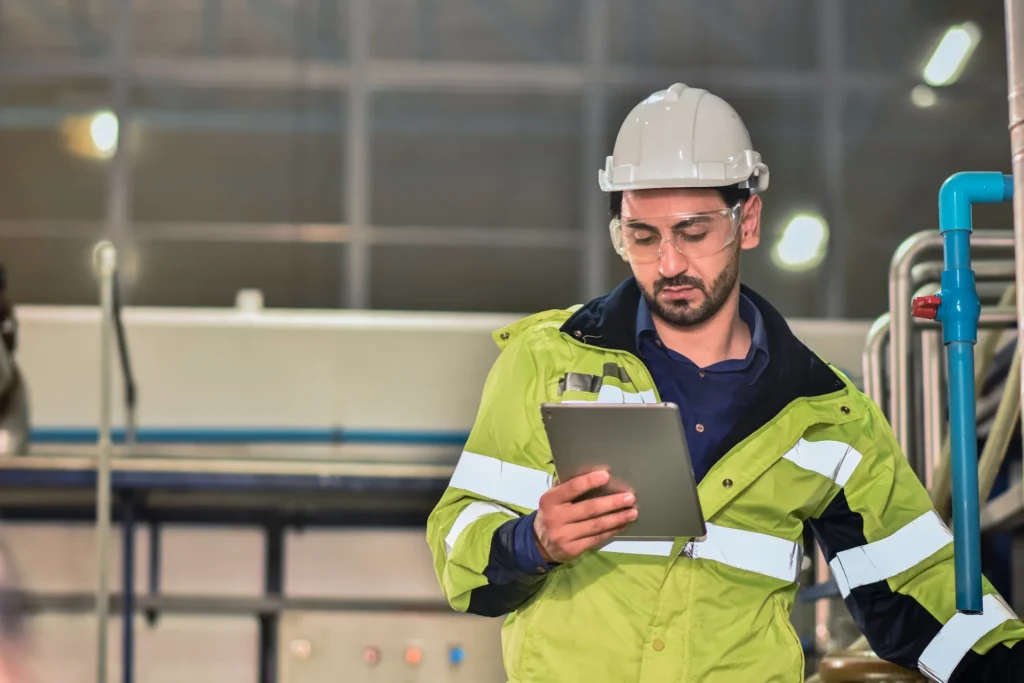 Industrial worker in a high-visibility jacket and hard hat examining a tablet in an industrial setting with safety glasses on.