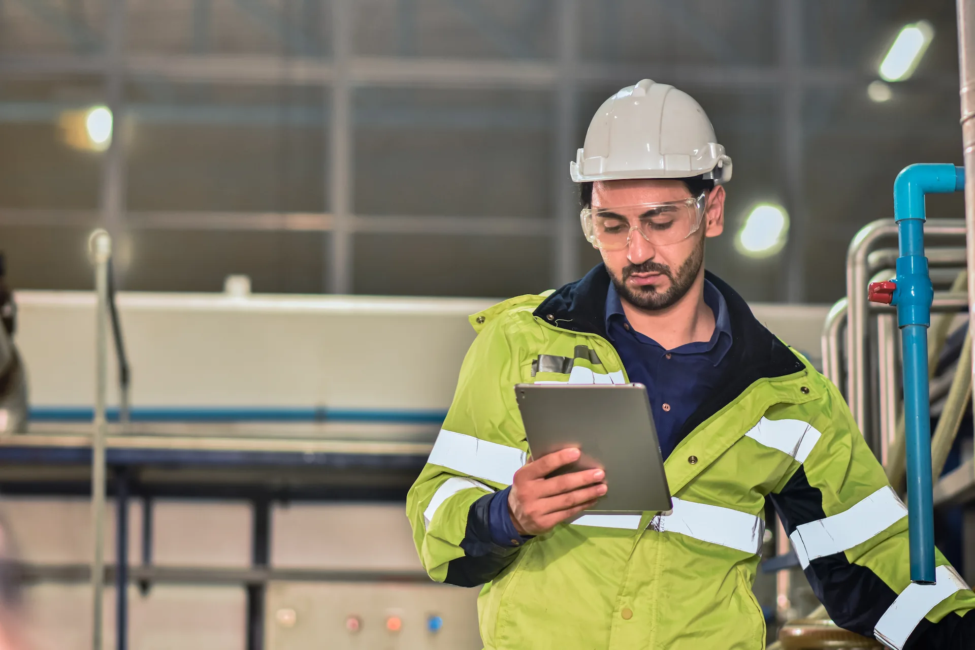 Industrial worker in a high-visibility jacket and hard hat examining a tablet in an industrial setting with safety glasses on.