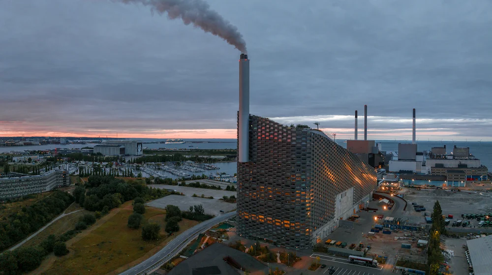 Aerial view of a large industrial power plant with a tall smokestack emitting dark smoke beside a waterfront at sunset, featuring a curved perforated building facade.Why it’s informative: identifies the power plant, smokestack, water setting, and time of day.